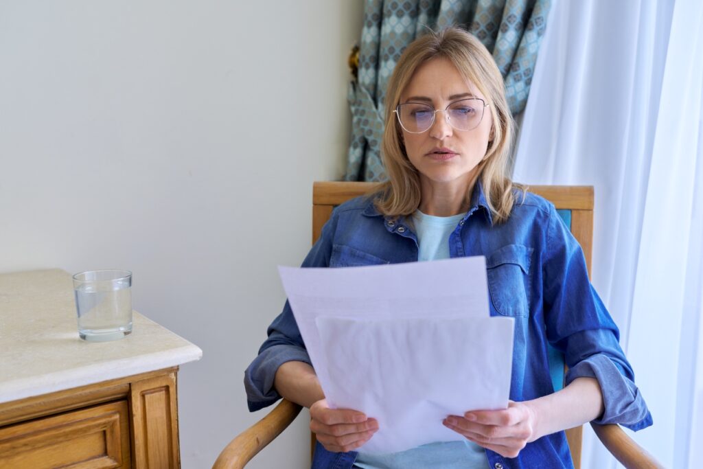 Serious middle aged woman with glasses reading papers sitting at home in a chair. Important business information, letter, prenup documents, lifestyle