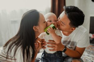 A mother and father kiss their baby’s cheeks while the child holds a small green toy, creating a warm and affectionate family moment indoors.